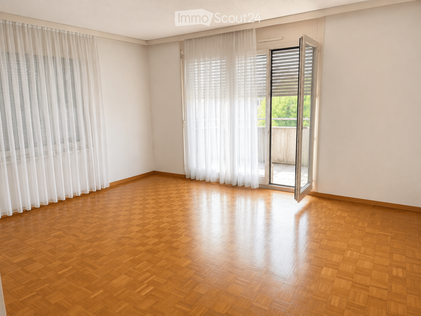 Empty room with wooden parquet flooring, white walls, large windows with sheer curtains, and a sliding glass door leading to a balcony.