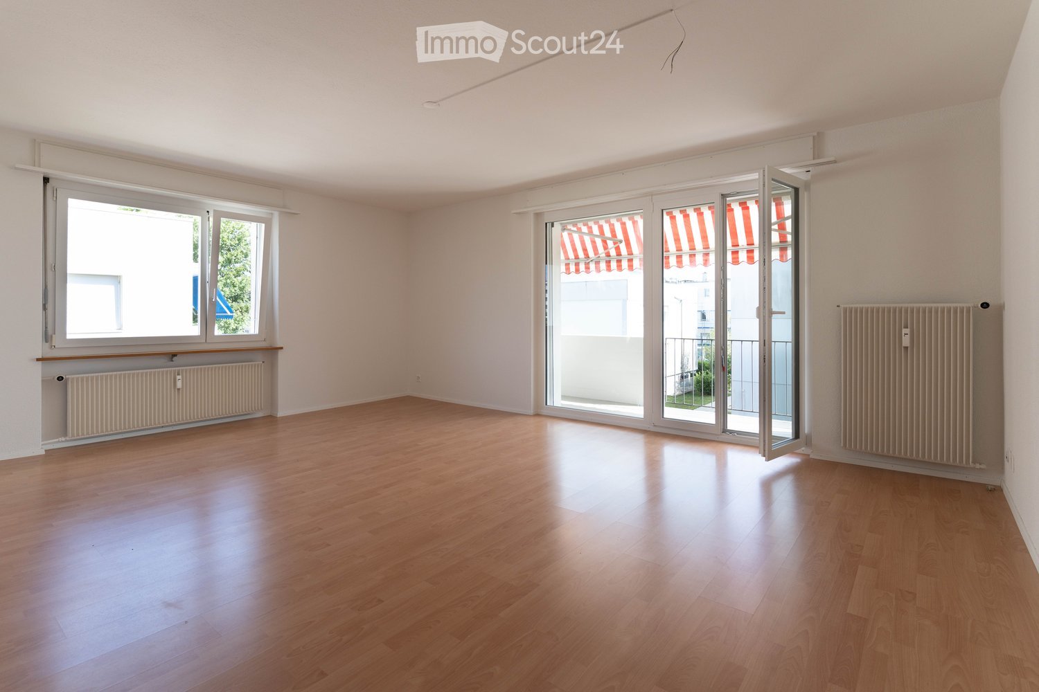 Empty room with wooden flooring, white walls, large windows, and sliding glass doors leading to a balcony area.