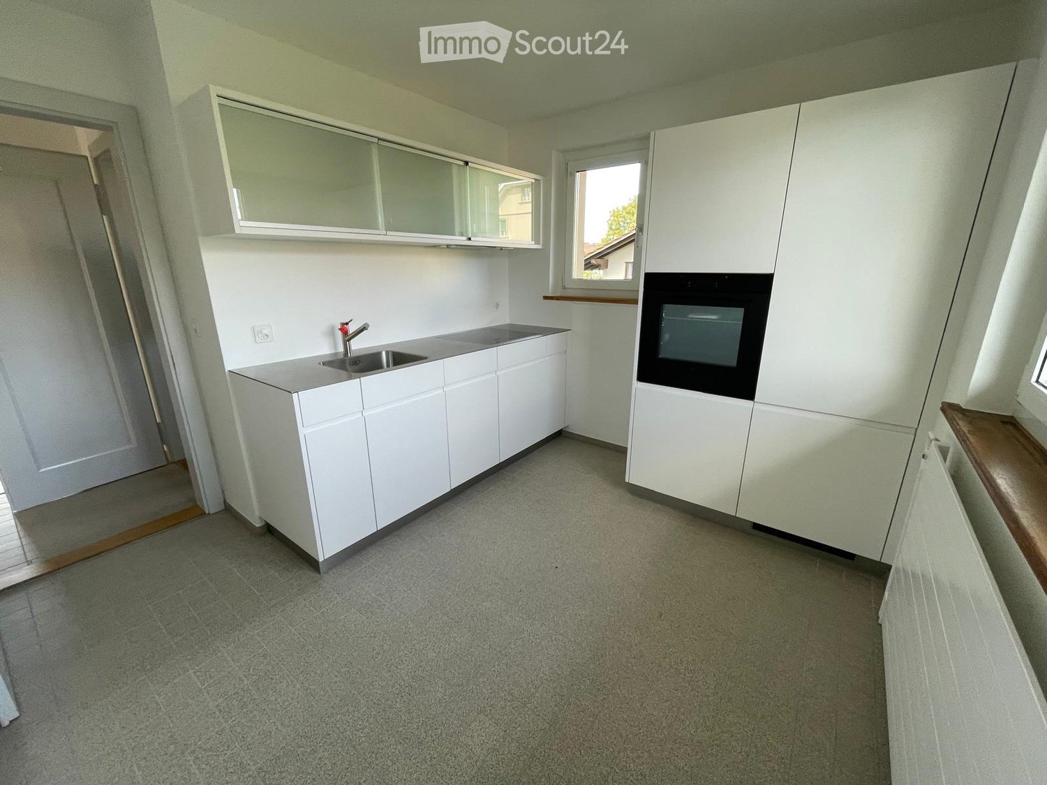 Empty kitchen with white cabinets, a sink, and a window, featuring a grey carpeted floor and a small wooden bench.
