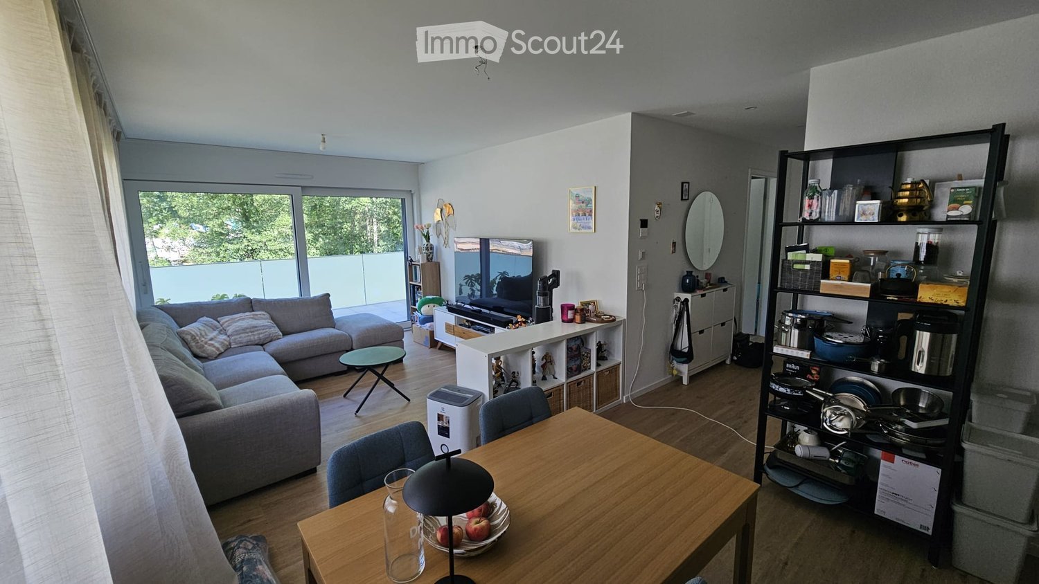 A modern, open-plan living and dining area with a grey sectional sofa, a wooden dining table with chairs, a small round coffee table, a TV stand, and a tall shelving unit holding kitchen items and decorative objects.