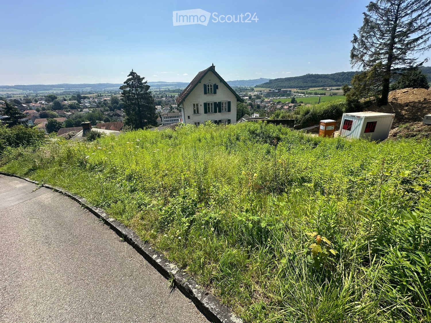 A grassy hillside with a paved pathway leading to a view of a town and surrounding landscape, with trees and a house visible in the distance.