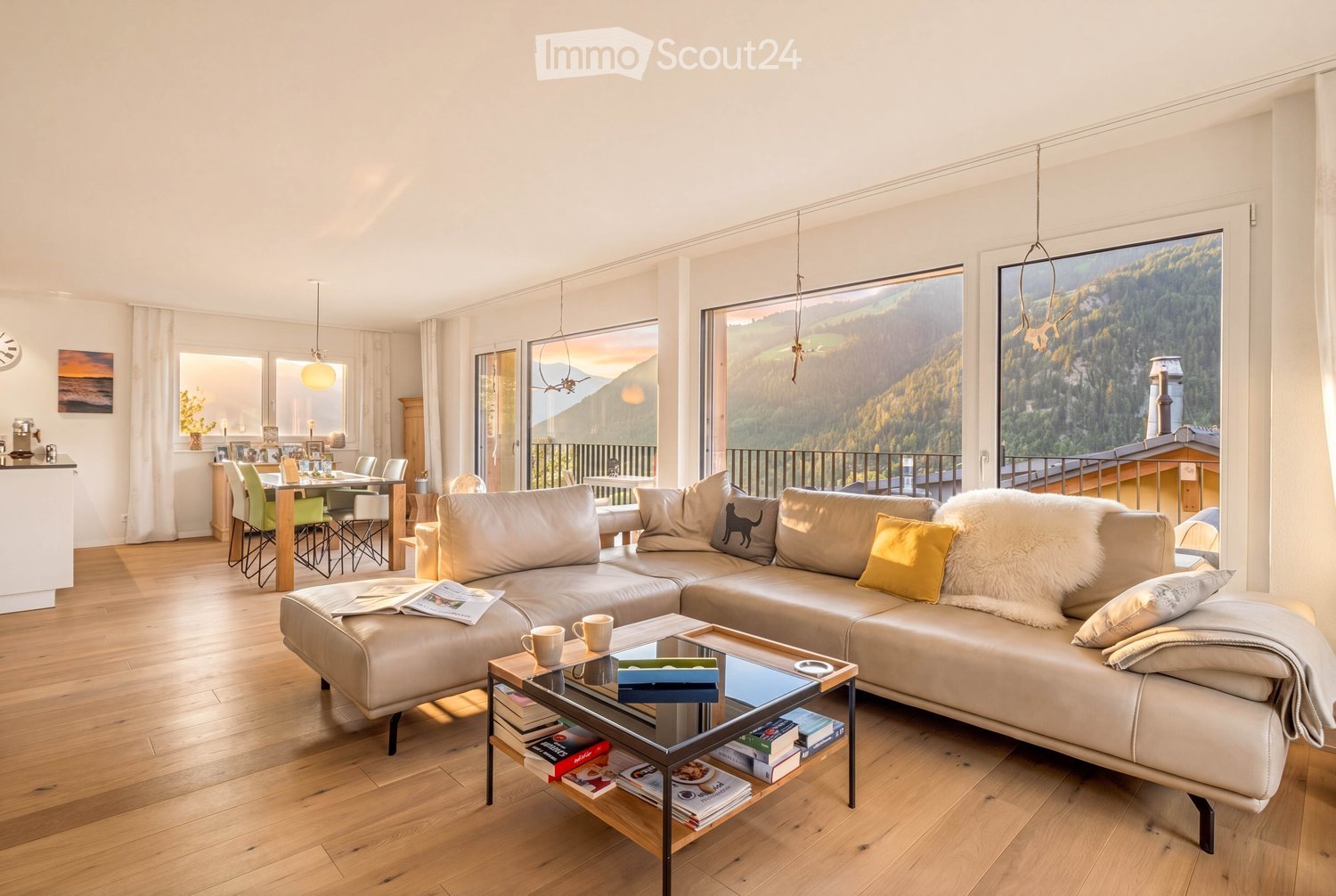 Bright living room with large windows offering a scenic mountain view, featuring a cream-colored sectional sofa, a glass coffee table with books and cups, a dining area with chairs and a table, and modern decor.