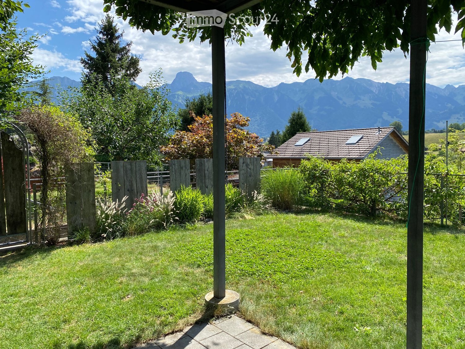 Lush green garden with a wooden fence, various plants, and a view of mountains in the background.