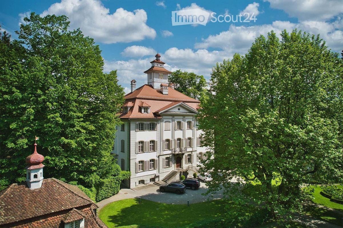 A large, elegant white mansion with a red-tiled roof, surrounded by lush greenery and trees. The mansion features multiple windows, a central entrance with steps, and a small tower with a dome on the roof. Several cars are parked in the driveway.