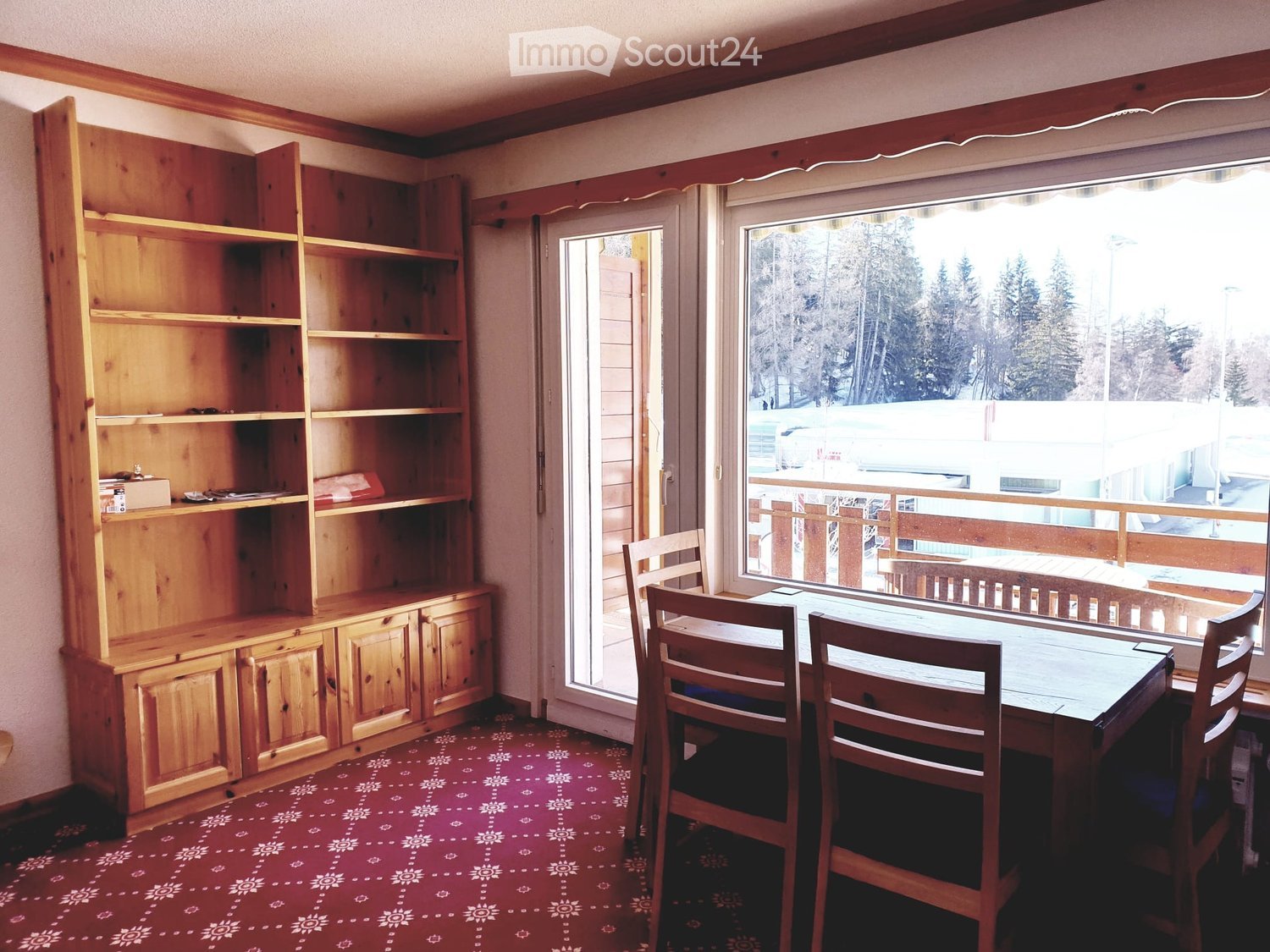 Dining area with a wooden table, chairs, and a wooden cabinet with shelves and drawers, overlooking a snowy outdoor scene.