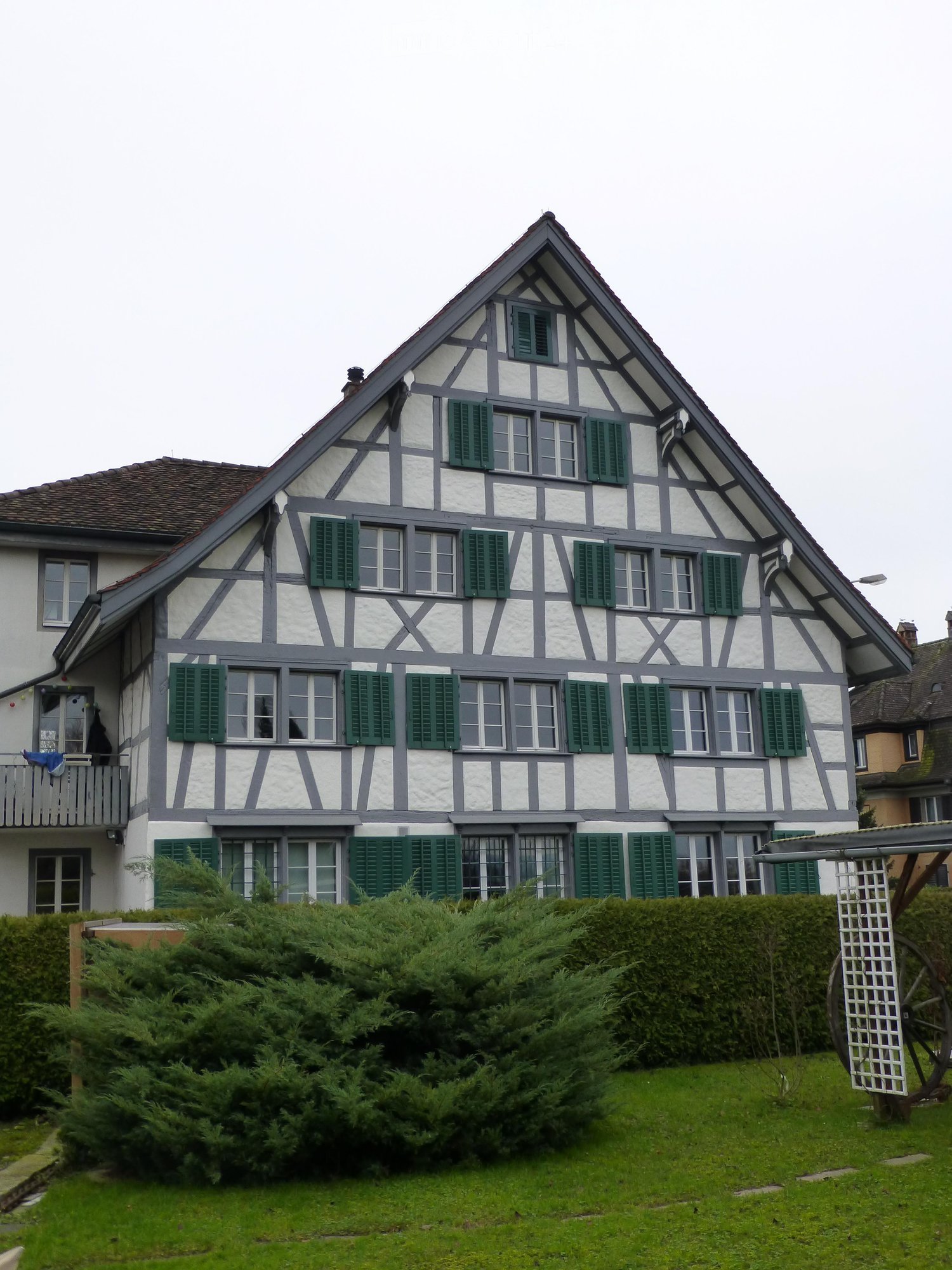 Two-story traditional half-timbered house with white plaster and dark wooden beams, green shutters, and a pitched roof.