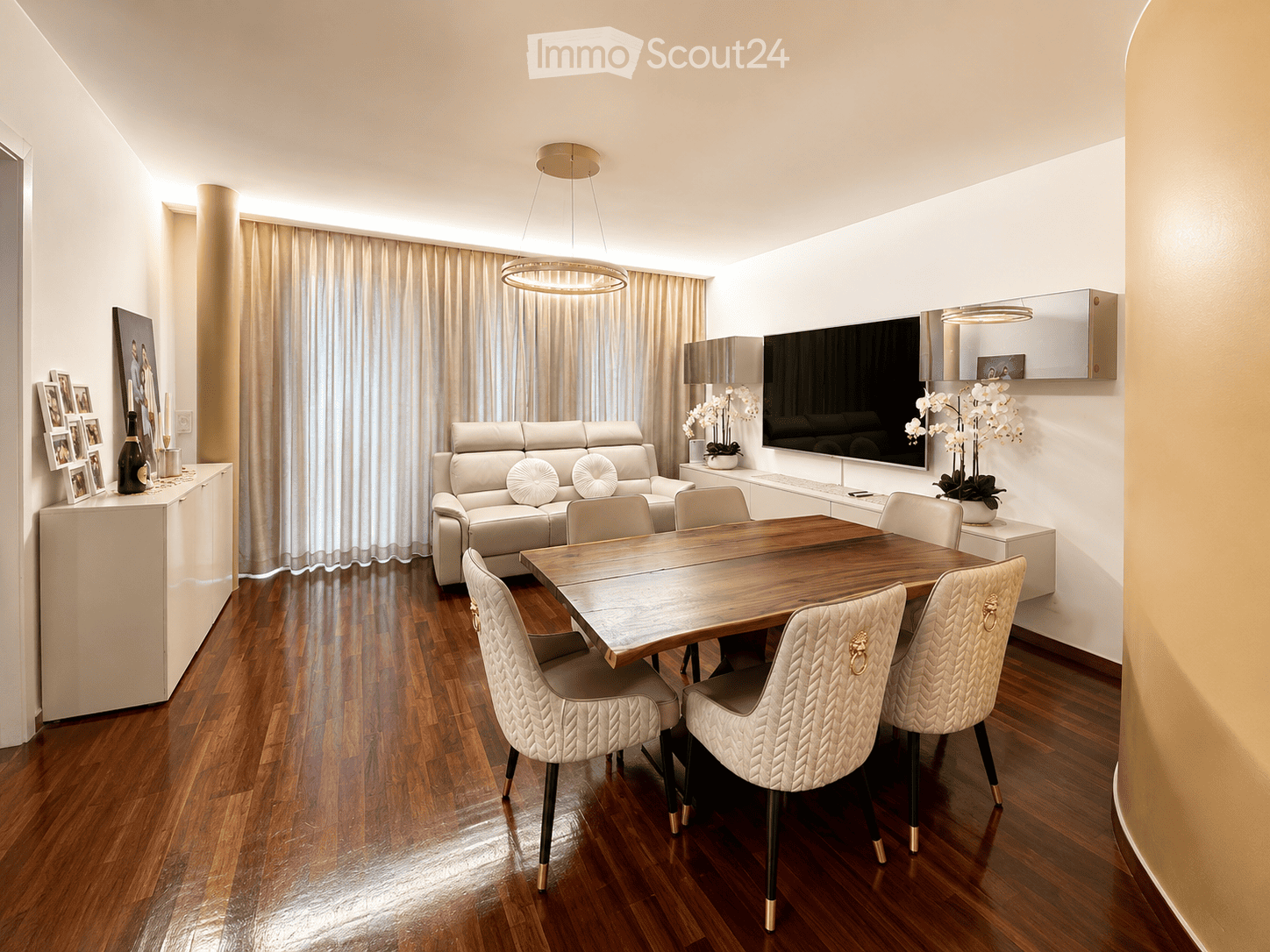 Elegant dining and living area with a wooden dining table, white upholstered chairs, a cream-colored sofa, a large flat-screen TV, and decorative elements including a flower vase and picture frames.