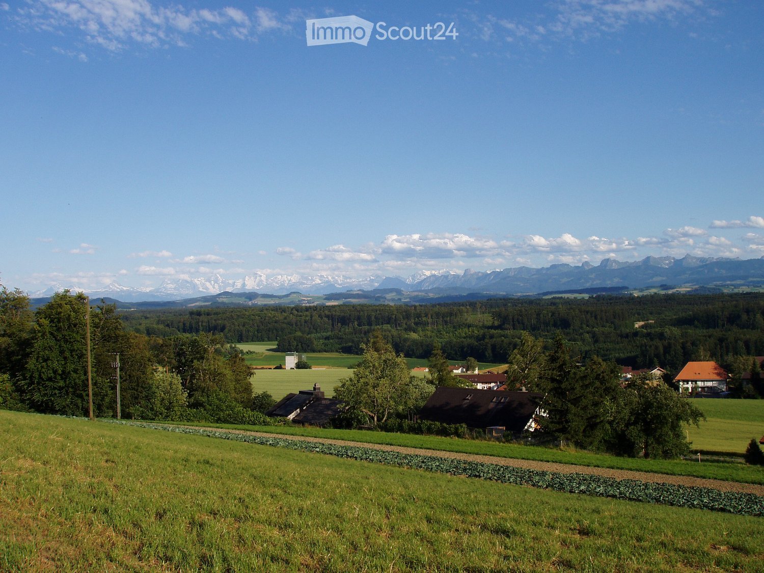 A rural landscape with green fields, scattered houses, and distant mountains under a clear blue sky.