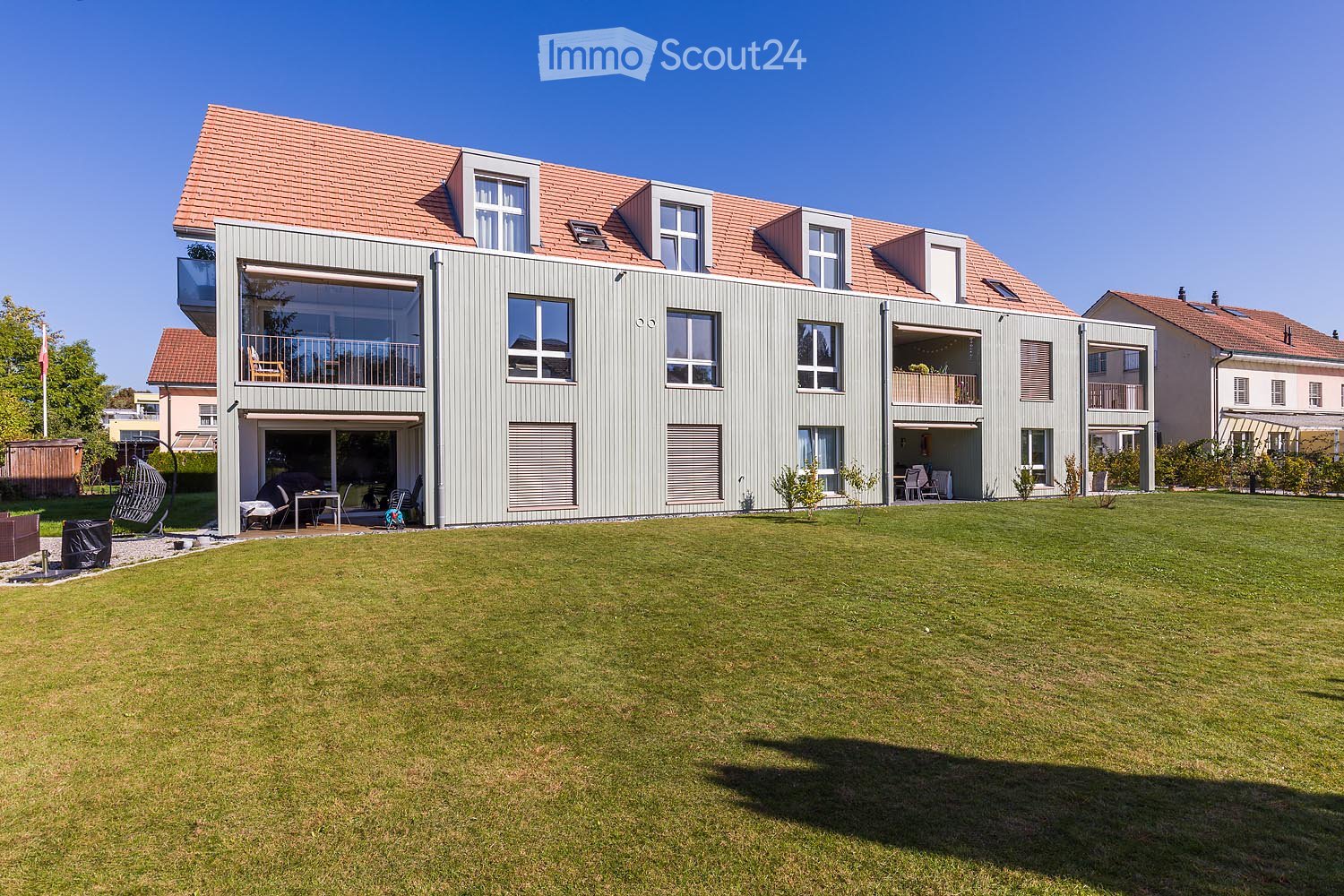 Modern two-story house with grey siding, red-tiled roof, multiple windows, balconies, and a spacious lawn.