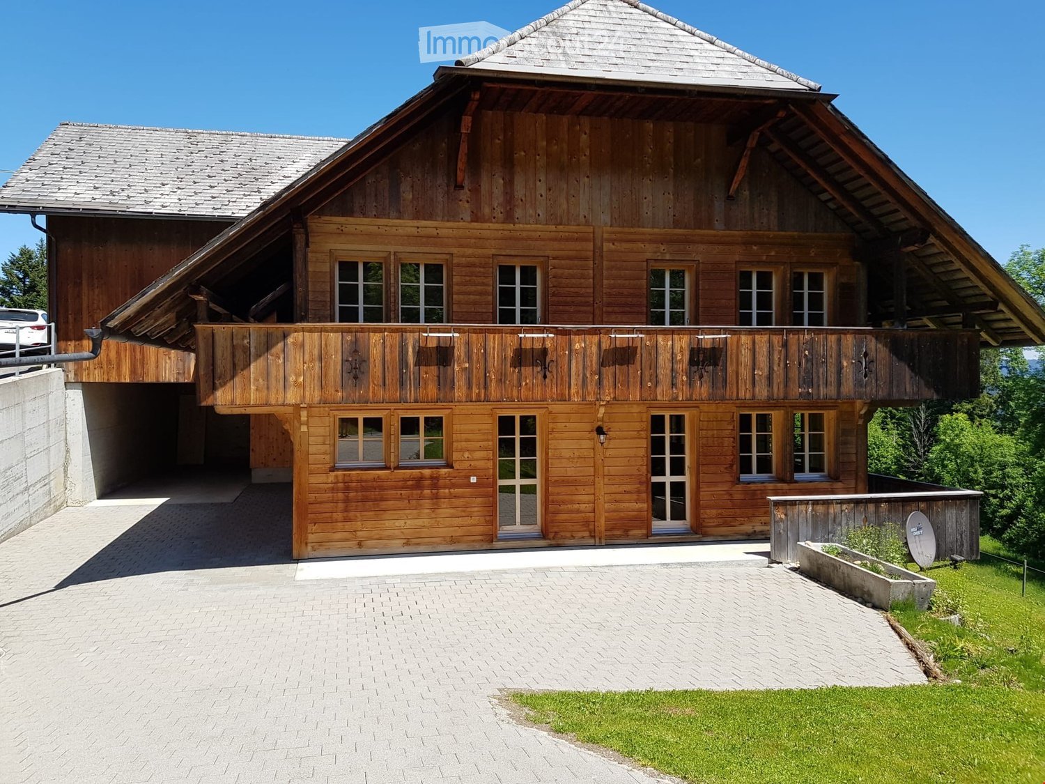 Two-story wooden house with a steep roof, large windows, and a wooden balcony. Features a paved driveway, garage, and small garden area.