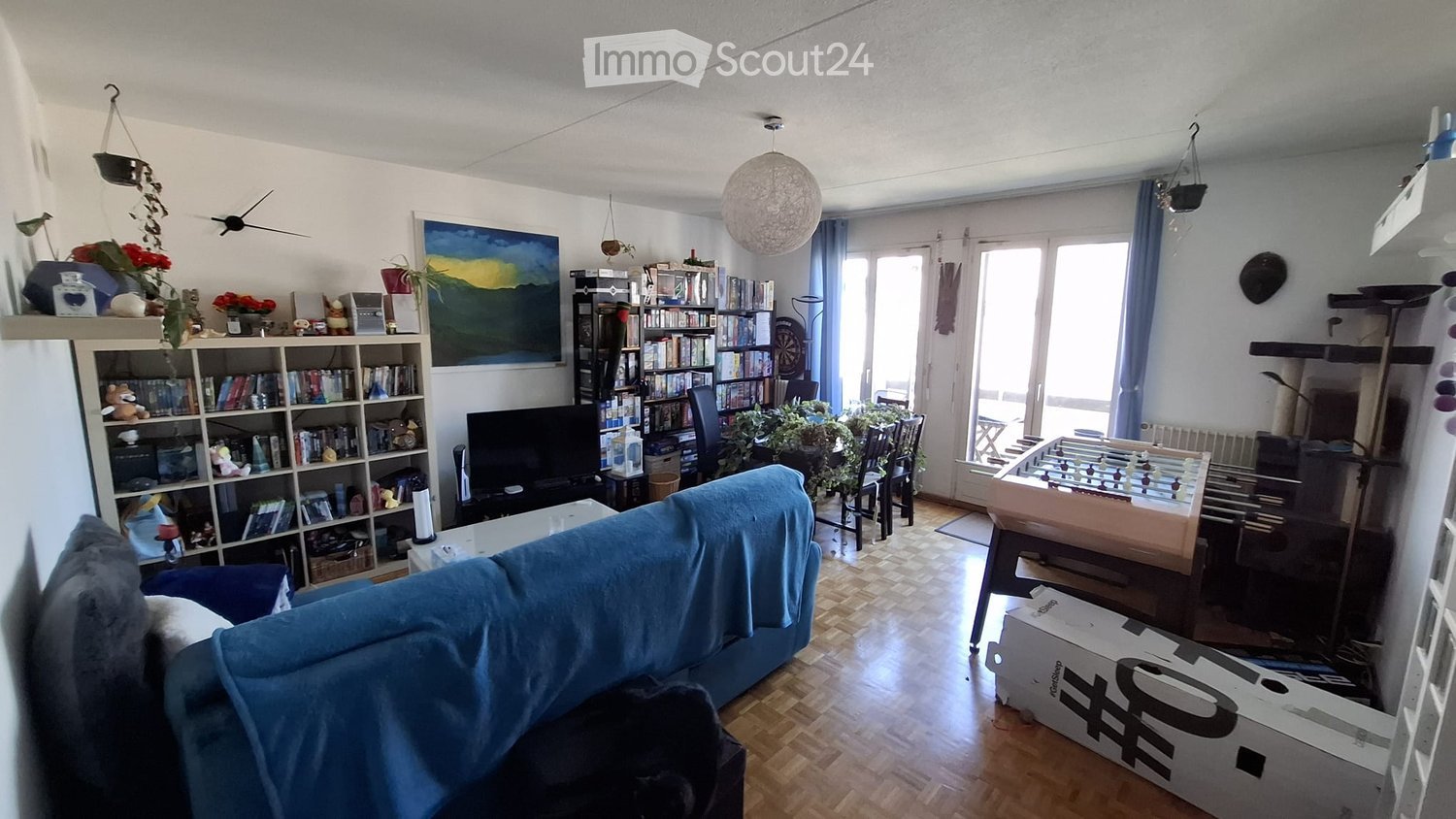 Living room with a sofa, coffee table, bookshelf, cat tree, football table, and a TV screen. The room has wooden flooring, large windows with curtains, and a painting on the wall.
