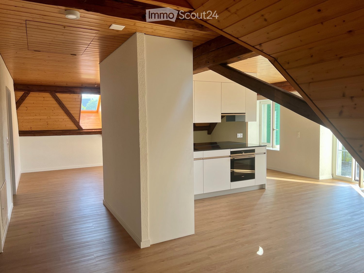 Open-plan attic space with wooden beams, light wood flooring, white cabinetry, and an integrated oven. Skylight and large windows provide natural light.