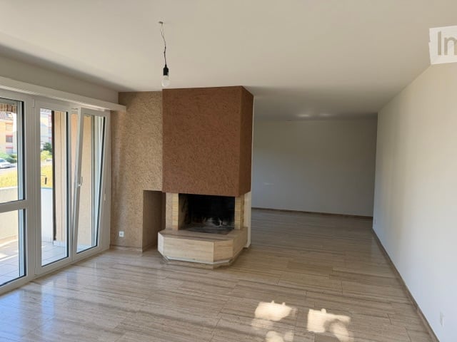 Empty room with wooden flooring, white walls, large glass doors, and a built-in fireplace with a stone base and wooden mantel.