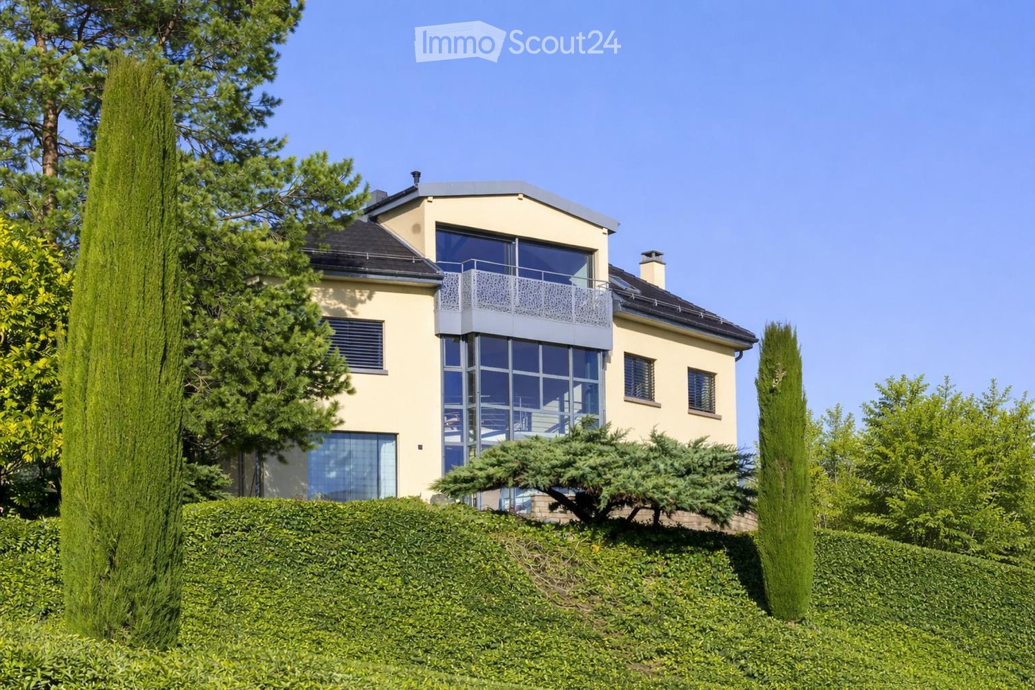 Two-story house with beige exterior, large windows, and a balcony on the upper level, surrounded by greenery and hedges.