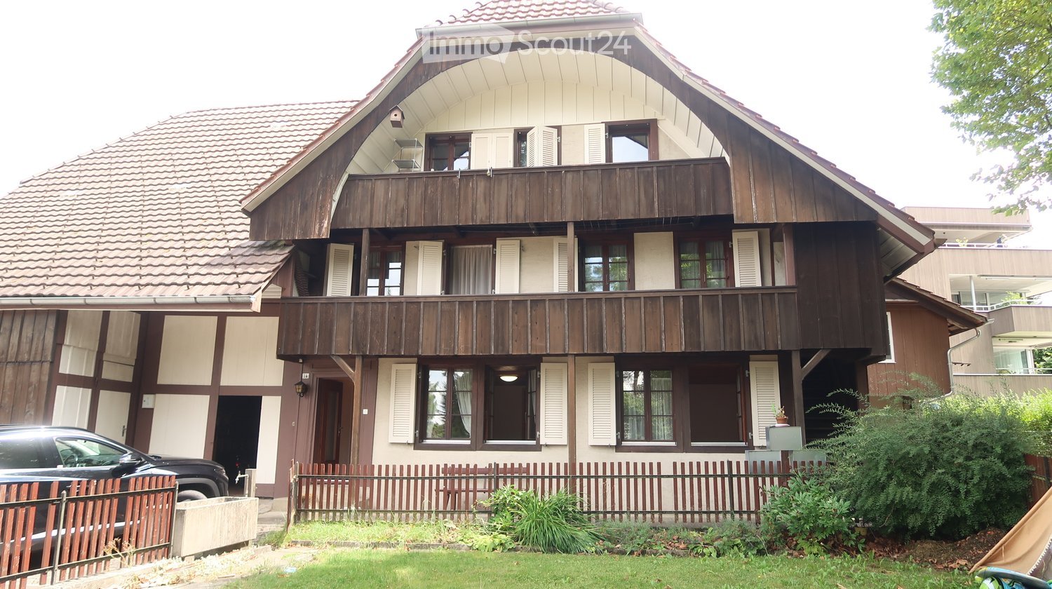 3 story house, brown roof, wooden exterior, balconies, black car parked in front, fence surrounding property, grassy area, potted plant, lamp over entrance