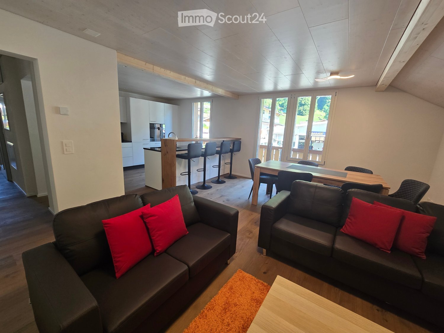 Modern living room with two black sofas, red cushions, a small wooden coffee table, a dining table with chairs, and a kitchen area with bar stools.