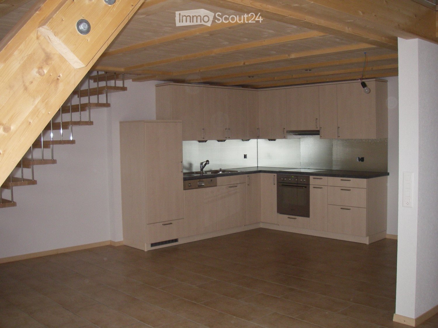 Modern kitchen with light wood cabinets, stainless steel sink, and built-in oven. The space features a tiled floor and a wooden staircase leading upwards.
