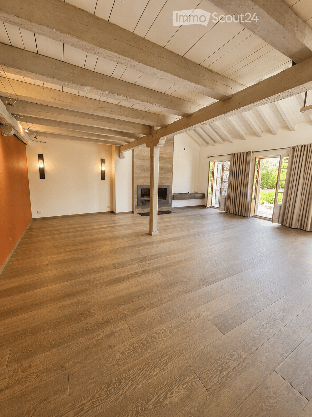 Spacious, unfurnished room with exposed wooden beams, hardwood flooring, and a fireplace. Large windows and sliding glass doors lead to an outdoor area.