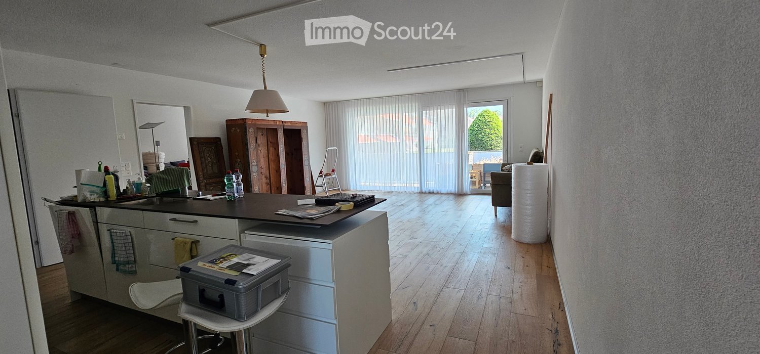 Open-plan kitchen and living area with wooden flooring, large windows, and a balcony. Kitchen island with stools, sofa, and various storage items.