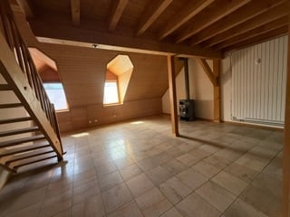 Empty attic space with wooden beams, tiled floor, and exposed brick walls. Features a wooden staircase leading up, a small window, and a radiator on the wall.
