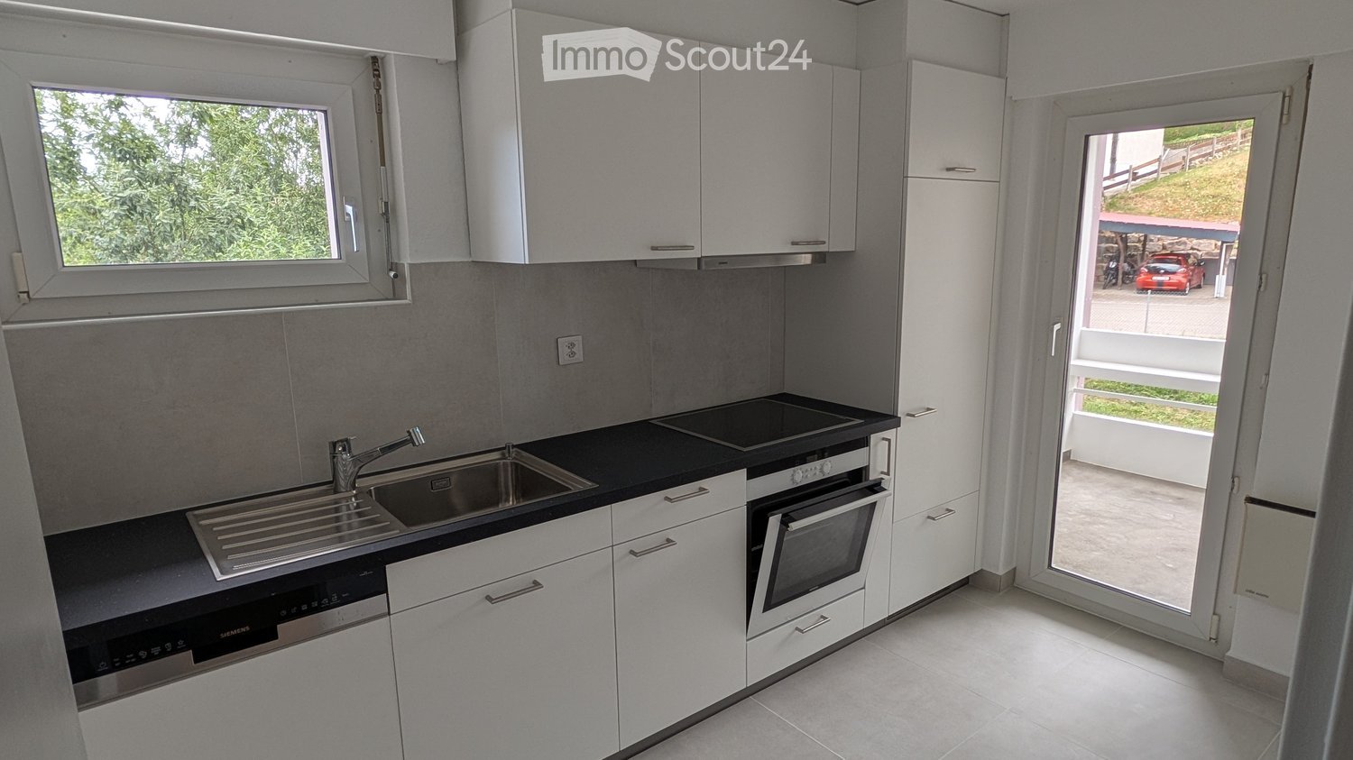 Modern kitchen with white cabinets, black countertop, stainless steel sink, oven, and dishwasher. Large window and sliding door leading to outdoor area.