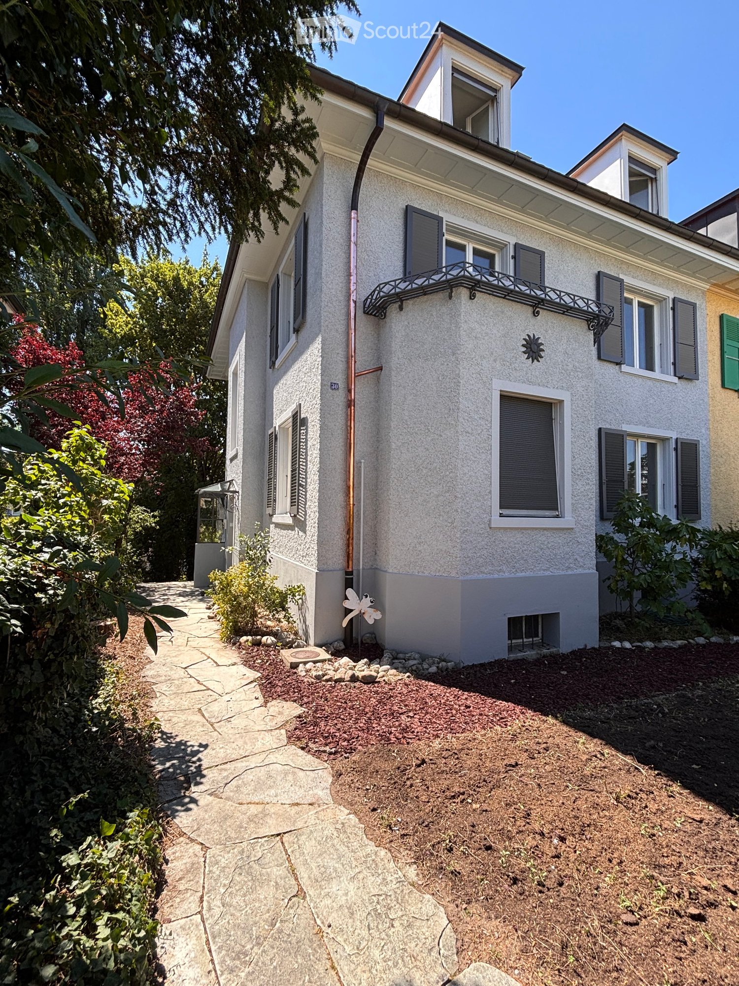 Two-story house with white facade, multiple windows, shutters, and a small balcony. The house is surrounded by a garden with a stone pathway and various plants.