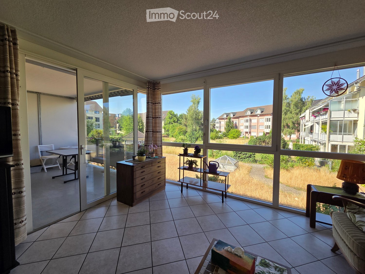 Bright living room with large windows offering a view of the neighborhood, furnished with a sofa, coffee table, chairs, a small table, a lamp, a cabinet, and potted plants.