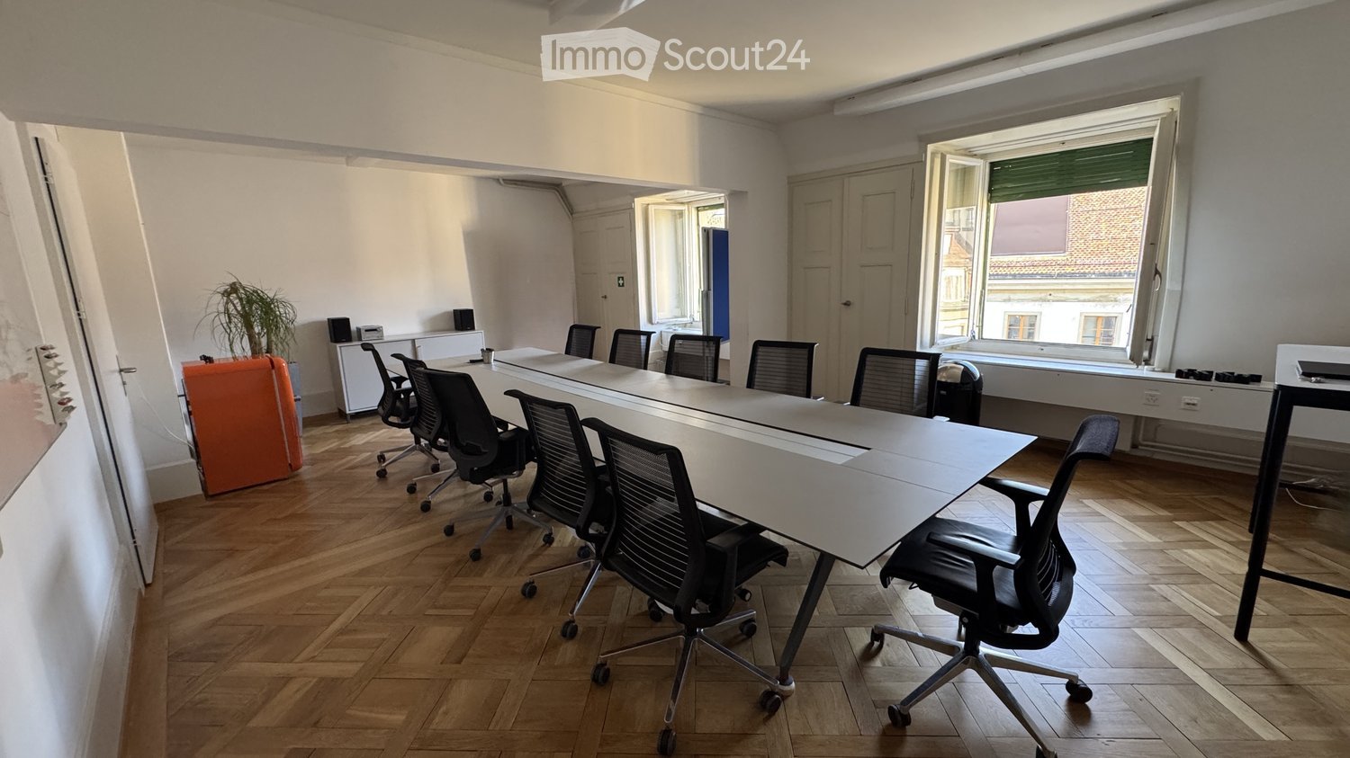 Bright conference room with a long white table, black office chairs, a potted plant, and a screen on the wall.