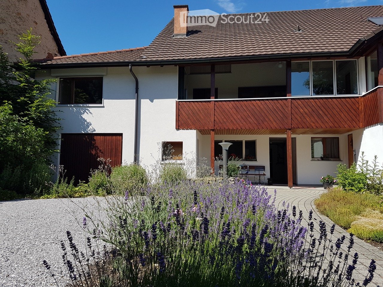 Two-story house with white exterior, wooden balcony, garage, and garden with lavender plants.