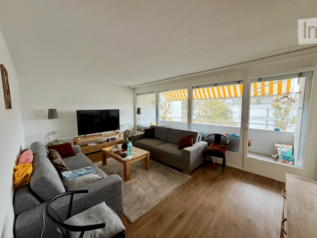 Bright living room with large windows and a sliding glass door leading to a balcony. Features a grey sectional sofa, coffee table, TV, and a small dining area with a chair.
