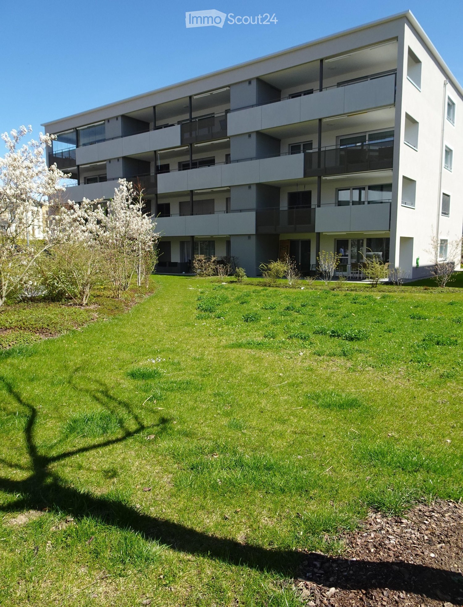 Modern multi-story residential building with balconies, surrounded by a well-maintained lawn and small garden area.