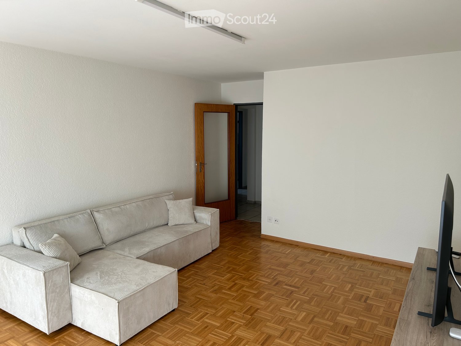 Modern living room with a grey sectional sofa, wooden flooring, and a flat-screen TV on a stand.