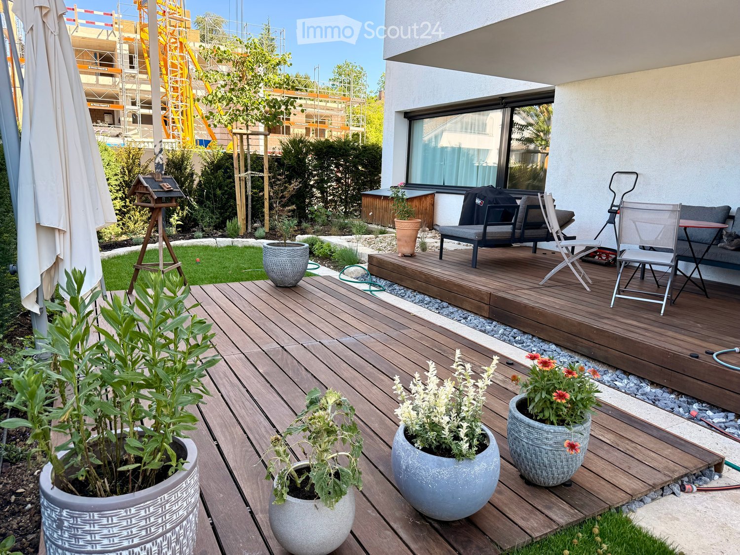 Wooden deck with potted plants, outdoor furniture, and a view of a garden area with a bird feeder and a small lawn.