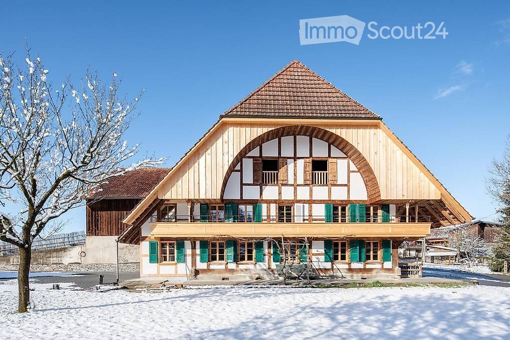Traditional wooden house with a steep roof, green shutters, and a balcony overlooking a snowy landscape.