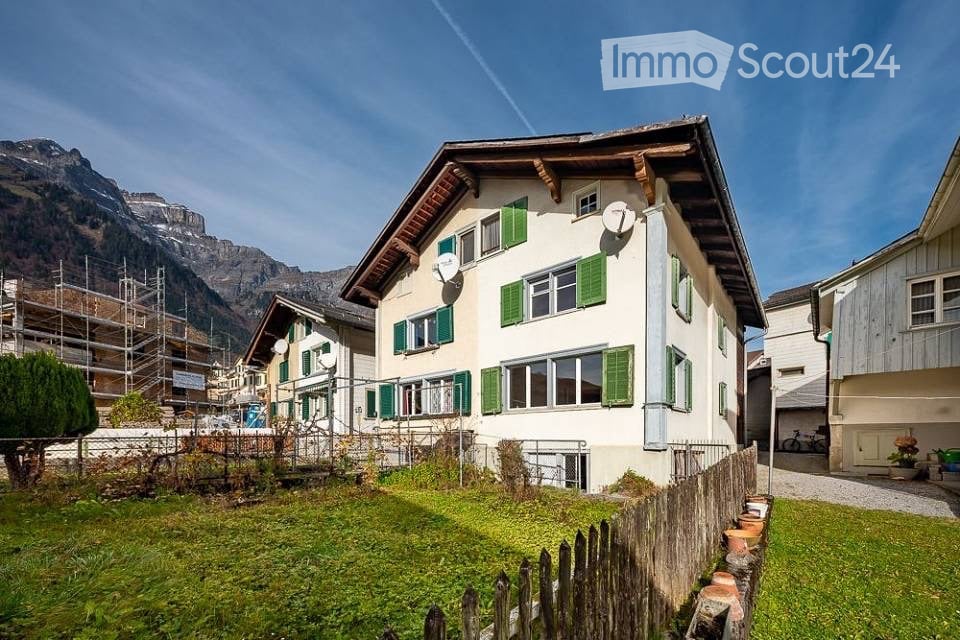 A two-story house with white walls, green shutters, and a wooden roof, situated in a scenic mountain village with a grassy front yard and wooden fence.