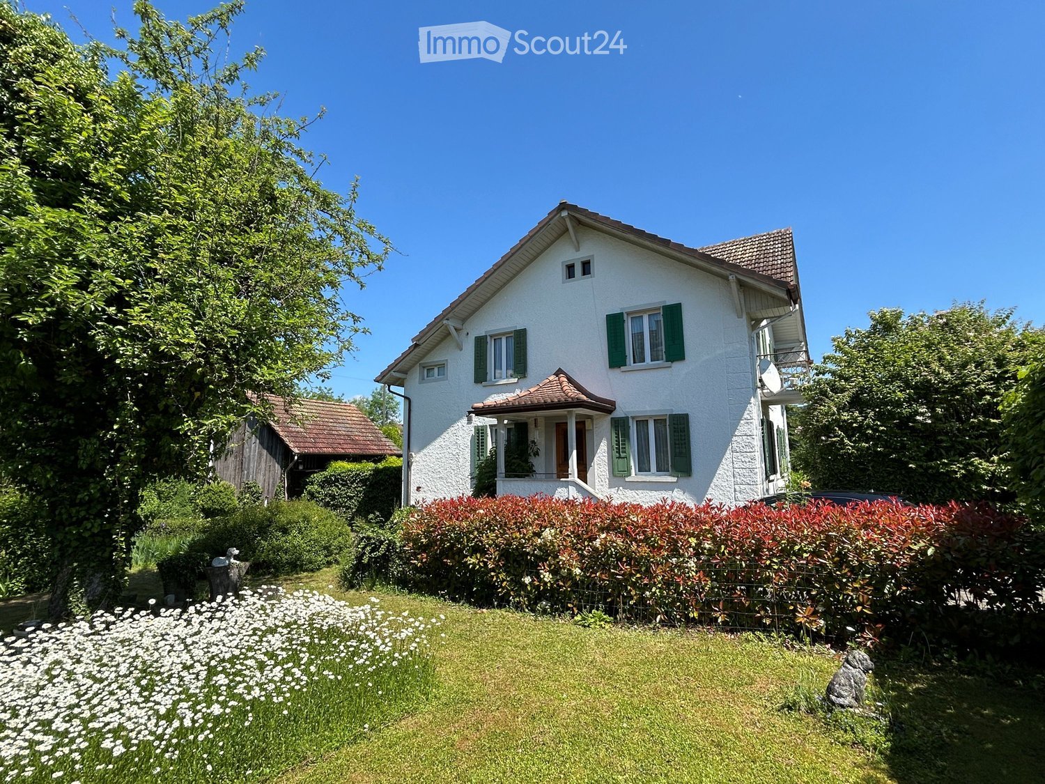 Two-story white house with green shutters, red-tiled roof, and a small balcony on the right side. The house is surrounded by a well-maintained garden with a hedge, flowers, and trees.