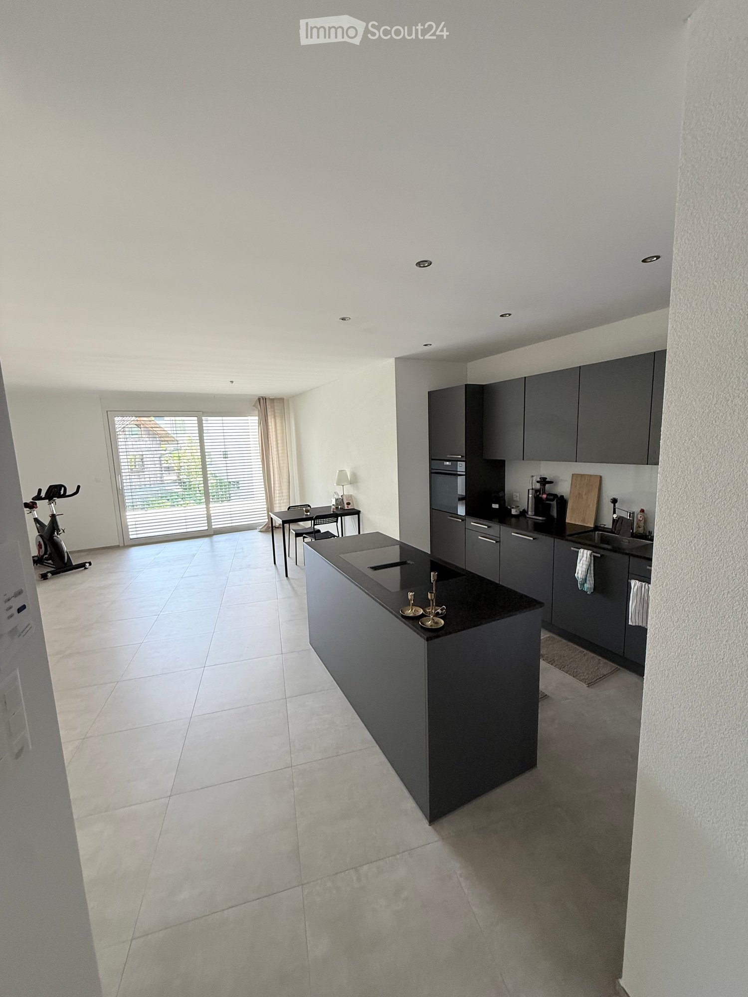 Modern kitchen with grey cabinets, a central island, and a dining area. Features include a sink, fridge, oven, and a small table with chairs.