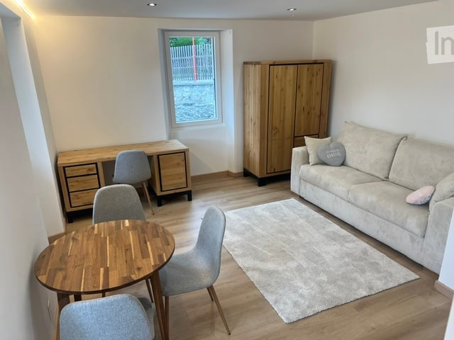 A modern, well-furnished room with a sofa, coffee table, chairs, a desk, a cabinet, and a small round table with chairs. The room features light wooden flooring and a neutral color palette.