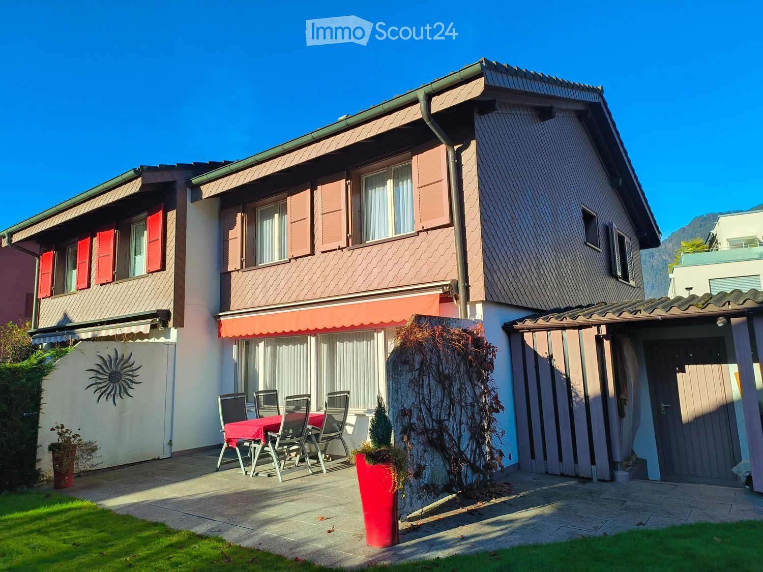 2 story house, brown exterior, red shutters, awning, outdoor dining set, red planter, garden, garage