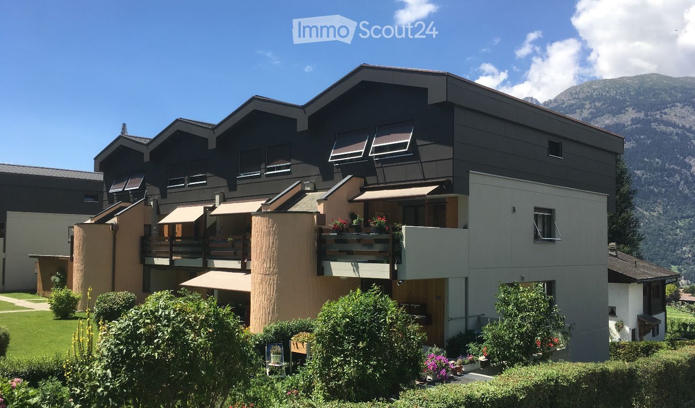 Modern multi-story building with balconies, surrounded by greenery and a scenic mountain view.