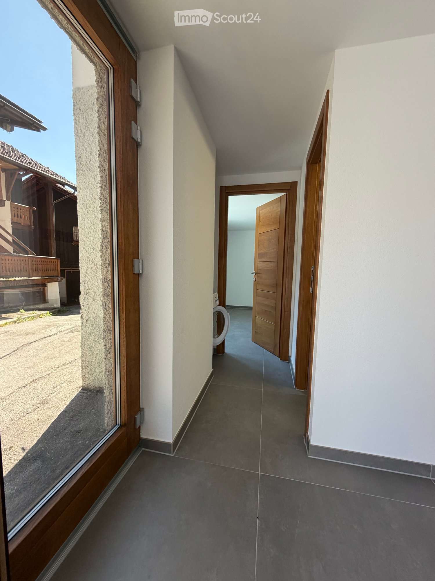 A narrow hallway with grey tiled flooring, white walls, and wooden doors leading to other rooms. A washing machine is visible in an adjacent room.