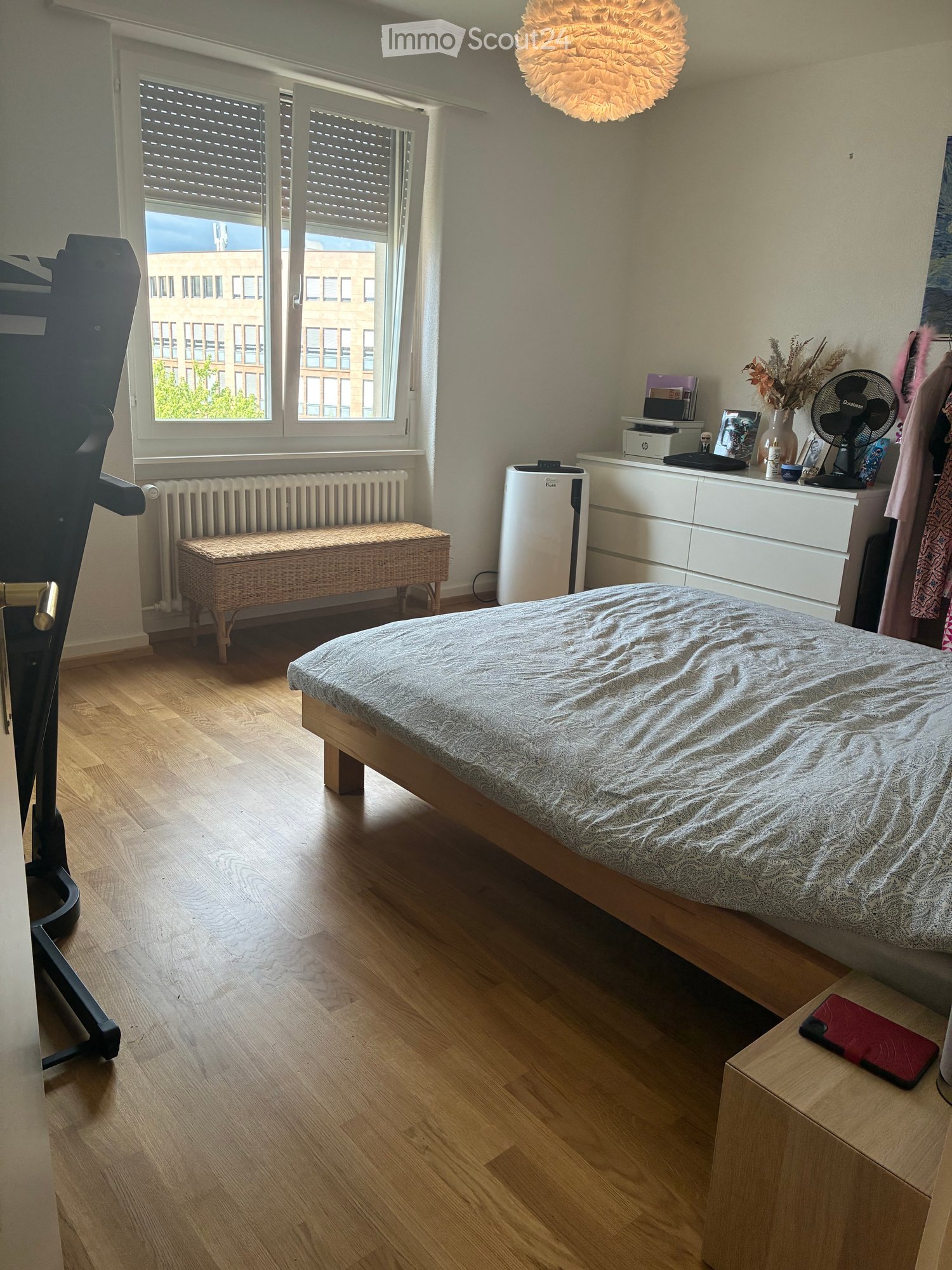 Cozy bedroom with wooden flooring, a double bed with patterned bedding, a nightstand with a fan, a radiator covered by a woven bench, and a white air purifier. The room features large windows with blinds and a modern pendant light.
