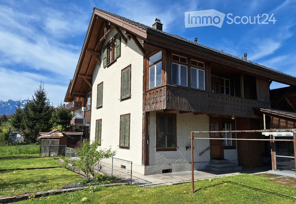Two-story house with white walls, green shutters, wooden balcony, and mountain view.