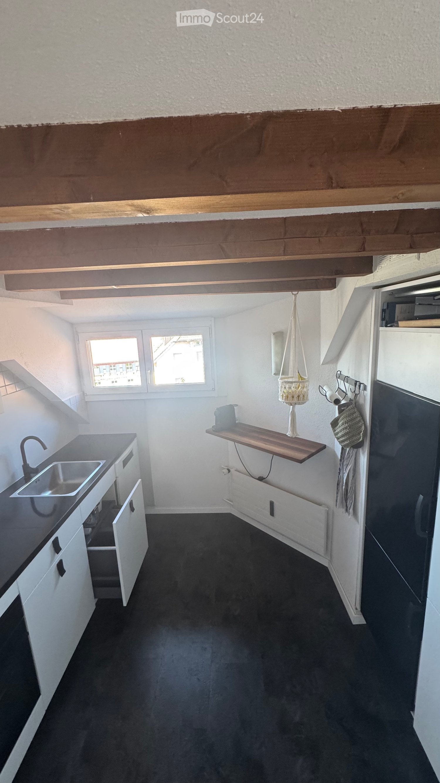 Modern kitchen with white cabinets, black countertop, stainless steel sink, and a wooden shelf with a hanging basket. The space features large windows and a dark floor.