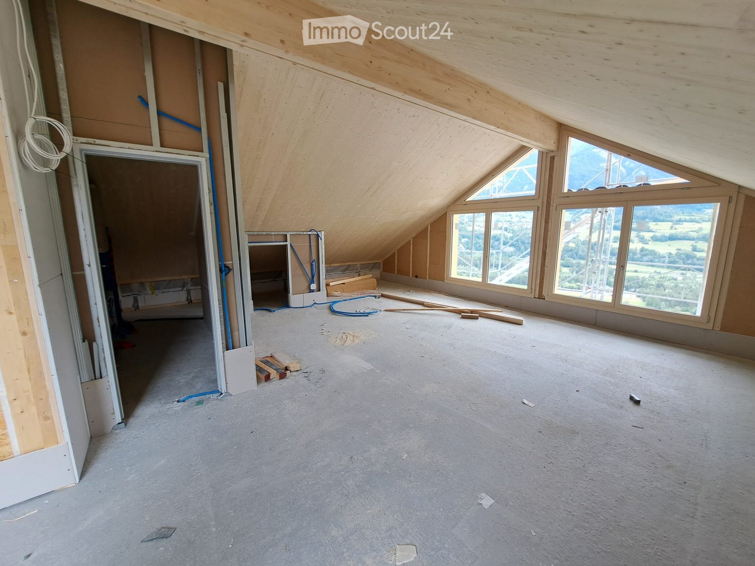 Empty attic space with exposed wooden beams, large windows offering a scenic view, and construction materials scattered around.