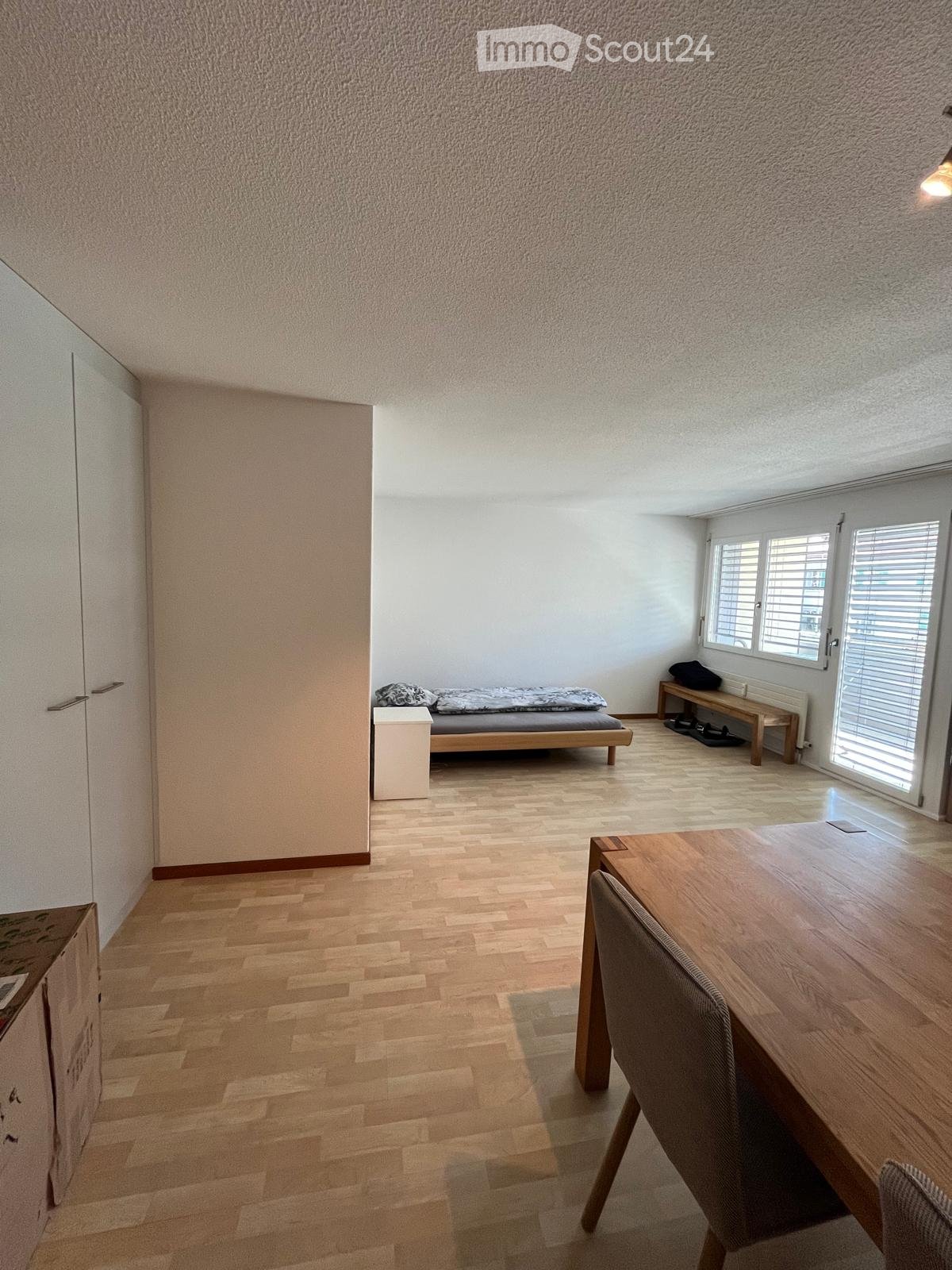 Bright, minimalist room with wooden flooring, a bed, a small table with a chair, and a bench near the window.