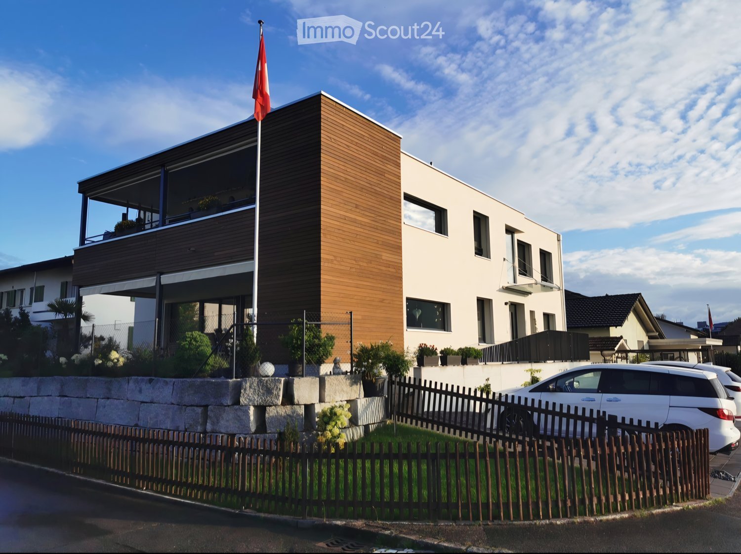 Modern two-story house with a combination of white and wood exterior, large windows, a balcony, and a flagpole with a flag.