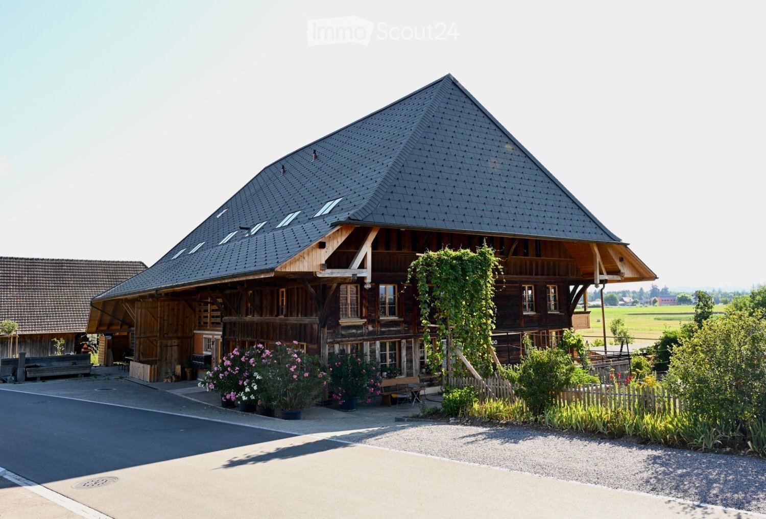 Traditional wooden house with a steep, dark roof and large windows, surrounded by a garden with flowers and a wooden bench.