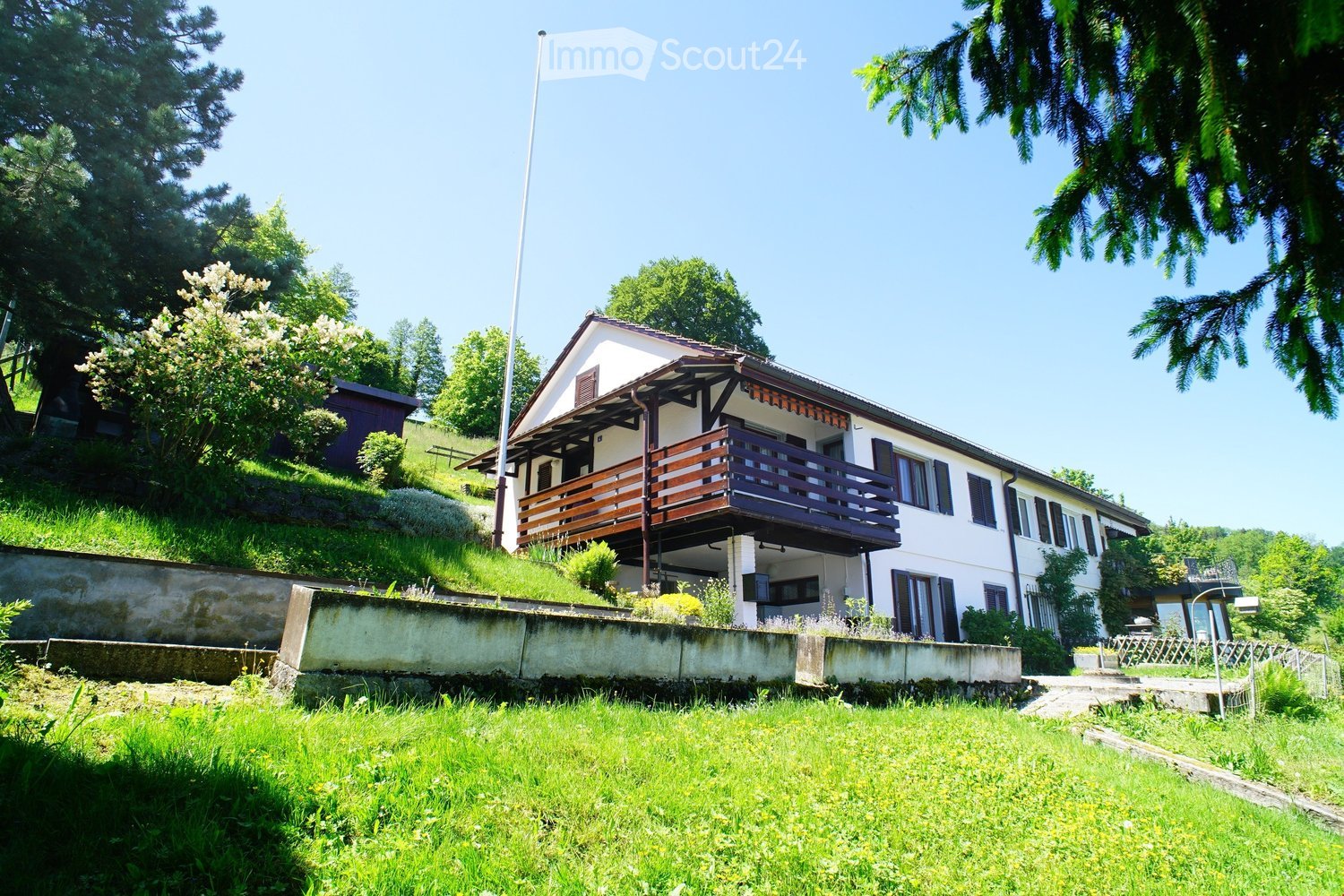 Two-story house with a raised ground floor, white exterior, wooden balcony, and surrounded by greenery.