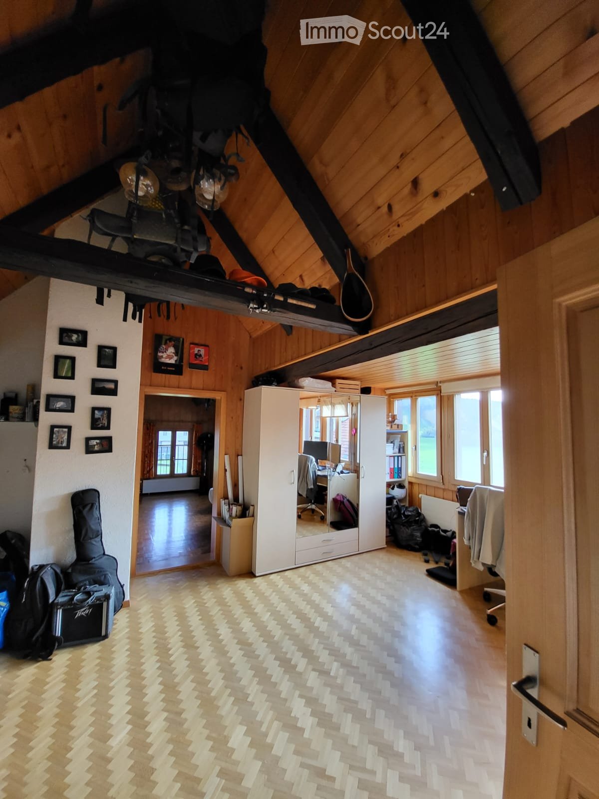 Spacious attic room with wooden beams, herringbone flooring, and large windows. Features a lofted sleeping area, a desk with a laptop, a large white cabinet with a mirror, and various personal items including a guitar and backpacks.
